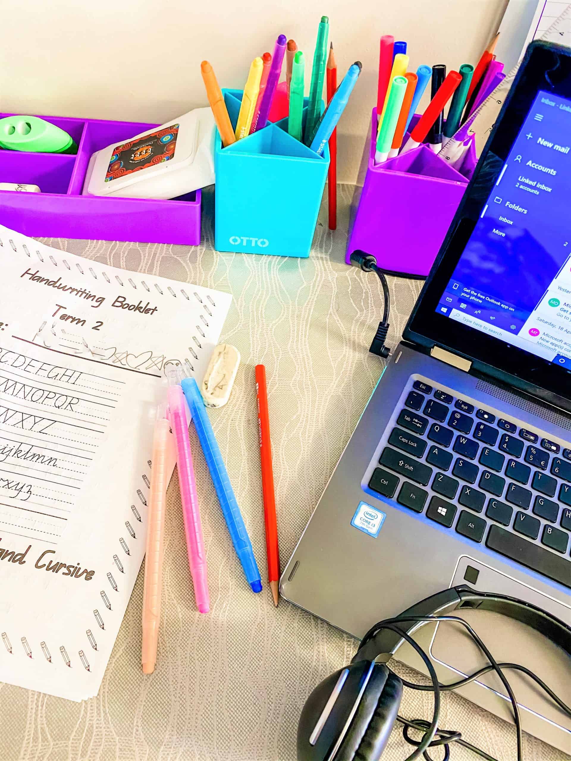 A desk with coloured pens, a handwriting booklet, rubber, a laptop showing a messaging app, and headphones on a light-patterned tablecloth.