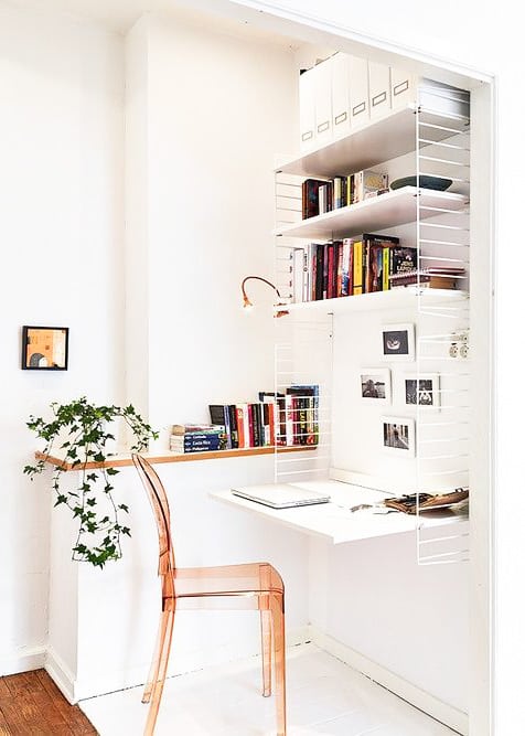 Small white home office nook with floating desk, wall shelves holding books and files, a transparent chair, potted ivy plant, wall lamp, and framed art on the wall.