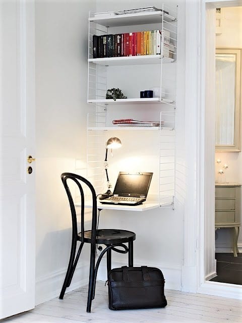 A small workspace with a black chair, wall-mounted shelves holding books, a laptop on a desk, a lamp, and a black briefcase on the floor in a white room.