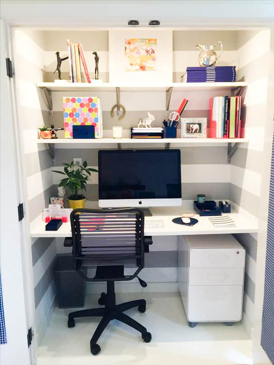Home office setup inside a small cupboard with a desk, black chair, iMac, office supplies, books, potted plant, and decorative items on shelves against striped walls.