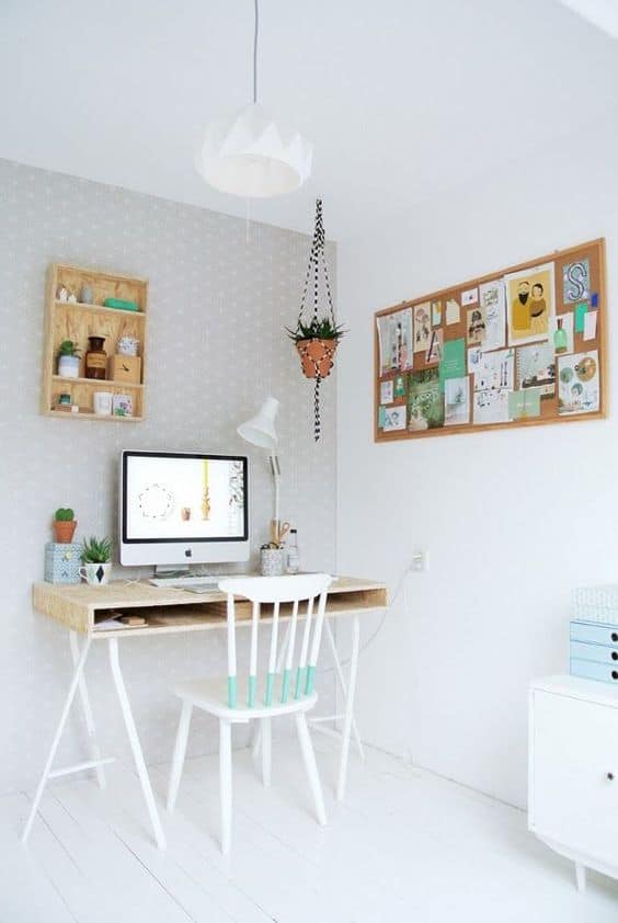 Minimalist home office with a wooden desk, white chair, desktop computer, lamp, wall shelves, hanging plant, and a corkboard with notes and pictures on a light grey and white background.