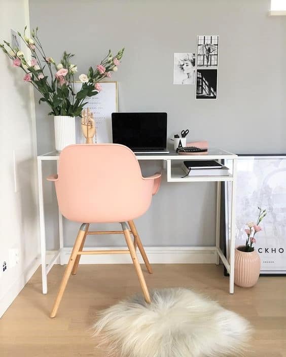 Minimalist desk setup with a pink chair, laptop, vase with flowers, framed art, and decor items on a light wood floor with a white fur rug.