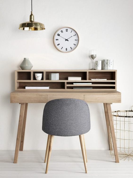 A wooden desk with shelves holds books and decor, paired with a grey chair. A round clock and pendant lamp hang on the white wall above.