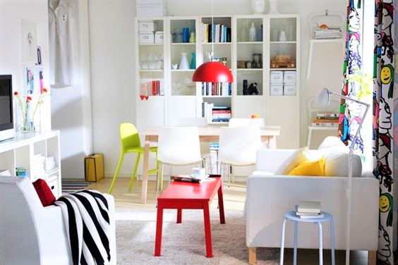 Bright living room with white furniture, a red coffee table, yellow and green chairs, bookshelves, and a red pendant lamp; colourful curtains add a playful touch.
