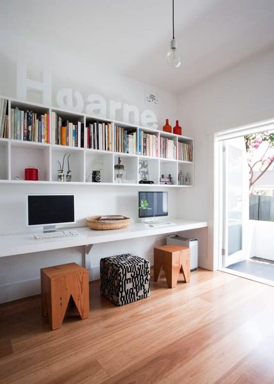 Minimalist home office with two computers on a white desk, wooden stools, patterned ottoman, wall shelves with books, and a door open to the outside.