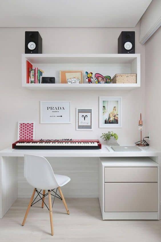A modern home office with a white desk, keyboard, chair, drawer unit, wall art, shelves holding books, decor, and speakers, and a small plant on the desk.