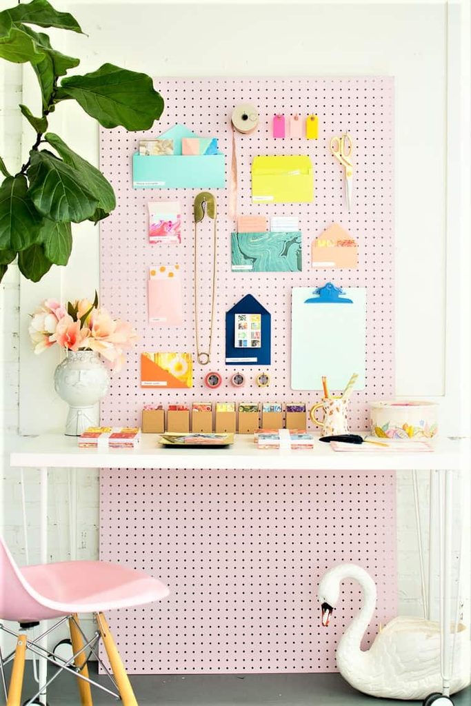 A white desk with a pink pegboard above it holds colourful office supplies and decorations; a pink chair and swan planter are nearby.