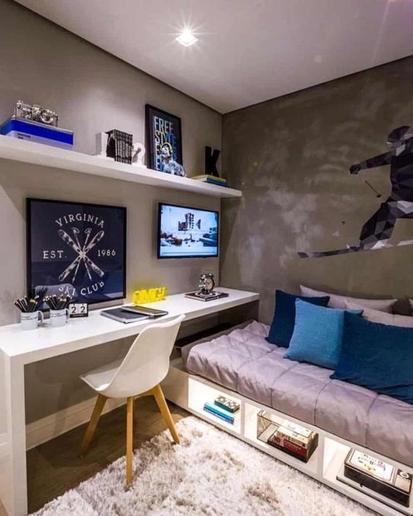 Modern bedroom with a bed, blue pillows, wall art, desk with chair, computer monitor, shelves, and books stored under the bed.