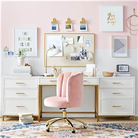 A modern white desk with gold accents, a pink velvet chair, wall art, photos, and office supplies, set against a pink wall.