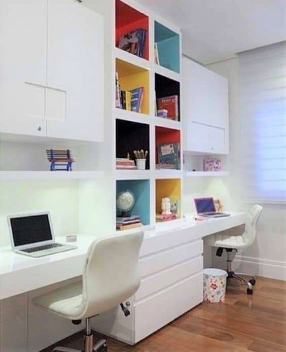 Two white desks with white chairs are set against a wall with colourful cubby shelves holding books and decor; laptops and office supplies are on the desks.