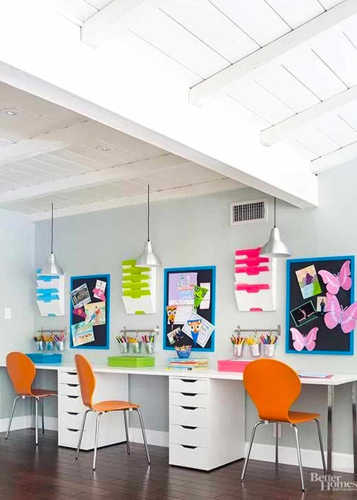 Three desks with orange chairs sit under pendant lights. Each desk has colourful art supplies, noticeboards with crafts, and drawers for storage against a light grey wall and white ceiling.