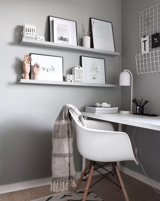 Minimalist home office with a white desk, lamp, and chair. Two wall shelves hold books, art, and décor. A checked blanket is draped over the chair, and a wire grid hangs on the wall.
