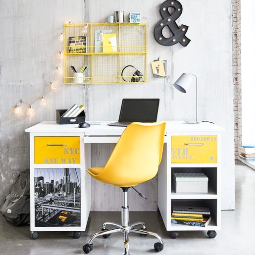 Modern desk setup with a yellow chair, laptop, lamp, wall shelf, decorative “&” sign, and NYC-themed decor against a grey wall.
