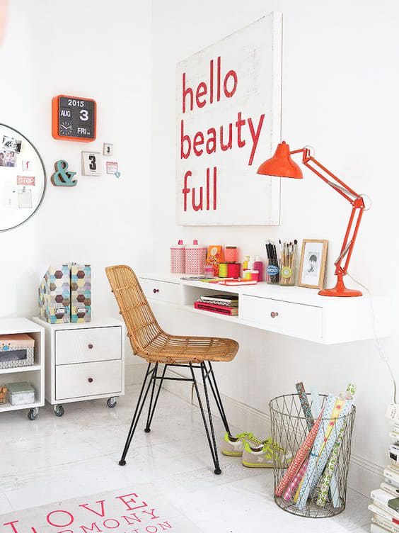 Bright, modern workspace with a wicker chair, floating desk, orange lamp, wall art reading “hello beauty full,” and colourful decor accents in a white room.