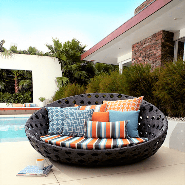 Round outdoor lounge chair with colourful patterned cushions placed on a patio near a pool, with greenery and a modern house in the background. A book and cup rest on the ground beside the chair.