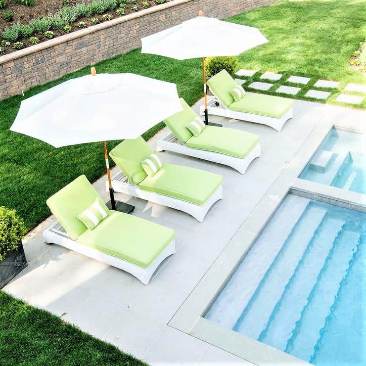 Four green loungers with white cushions and two white parasols are arranged beside a swimming pool on a stone terrace, with a grassy area and brick retaining wall in the background.
