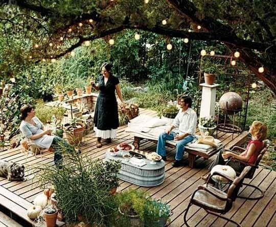 Four people sit and stand on a wooden decking surrounded by plants, food, and fairy lights, engaged in conversation in a lush outdoor garden setting.