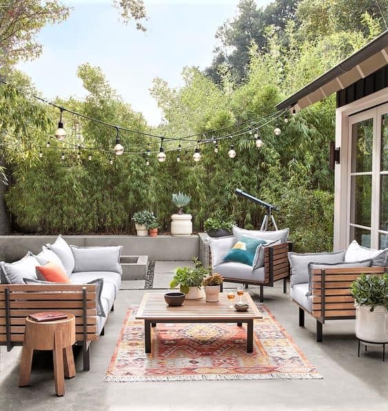 Outdoor patio with wooden sofas, a patterned rug, a coffee table, potted plants, fairy lights overhead, and a telescope near a wooden fence lined with tall bamboo.