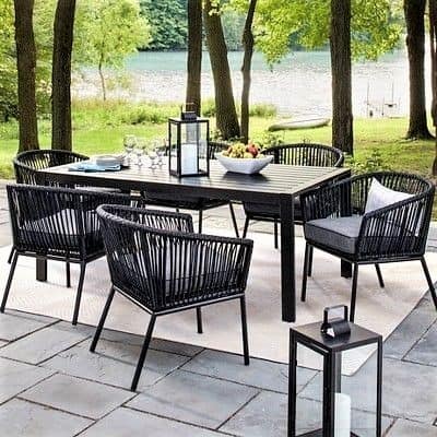 Outdoor patio set with black chairs and table, lanterns, and a fruit bowl on a stone floor, overlooking trees and a lake in the background.
