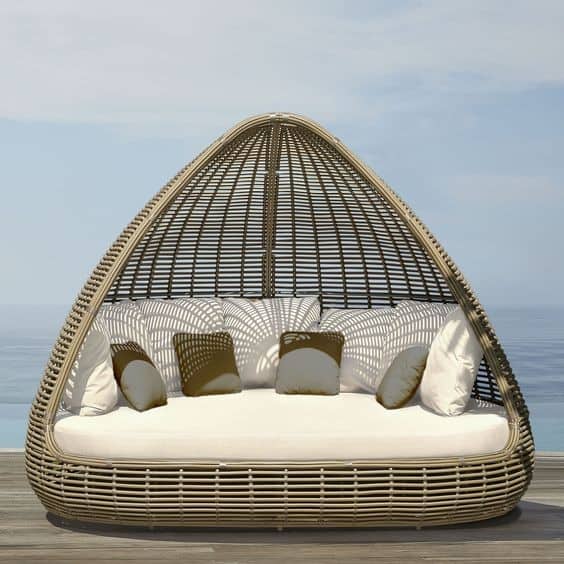 Wicker outdoor daybed with a dome-shaped canopy and multiple decorative cushions, positioned on a wooden decking with a sea view in the background.