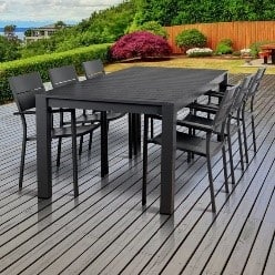 A black outdoor dining table with eight matching chairs on a wooden decking, surrounded by greenery and a wooden fence in the background.