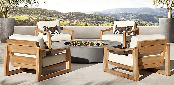 Four wooden armchairs with white cushions are arranged around a modern round firepit on a patio, overlooking a scenic landscape with hills and trees.