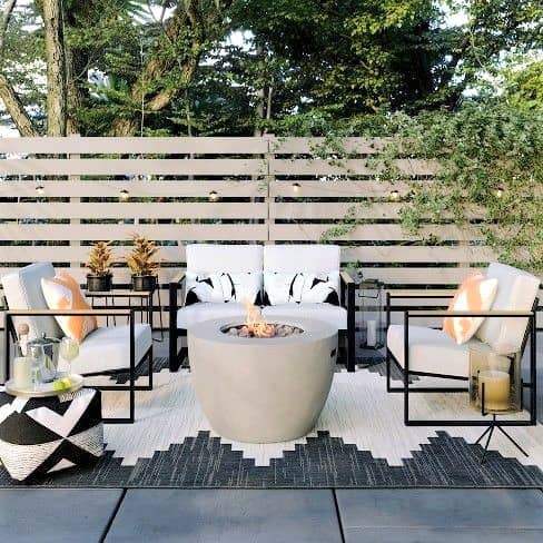 Modern outdoor patio with four cushioned chairs, a round fire pit in the centre, decorative cushions, and a patterned rug, set against a wooden privacy fence and surrounded by greenery.