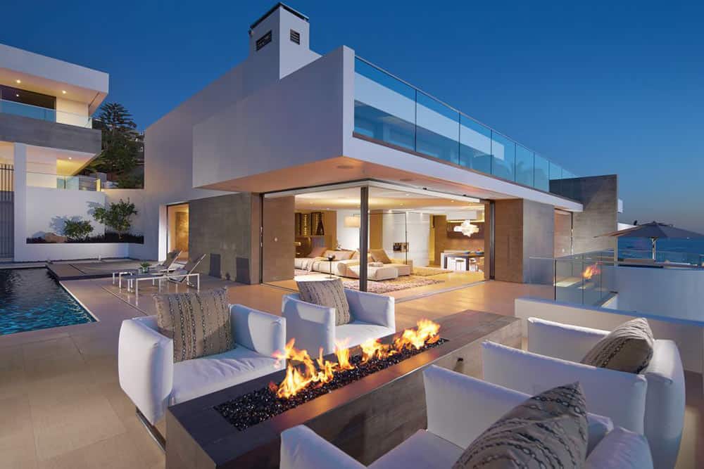 Modern luxury home with open living area, pool, and outdoor seating around a rectangular firepit at dusk.
