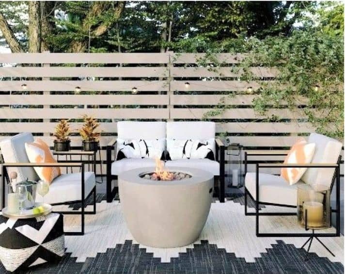 Modern outdoor patio with cushioned seating around a round fire pit, geometric rug, plants, and a wooden privacy fence in the background.