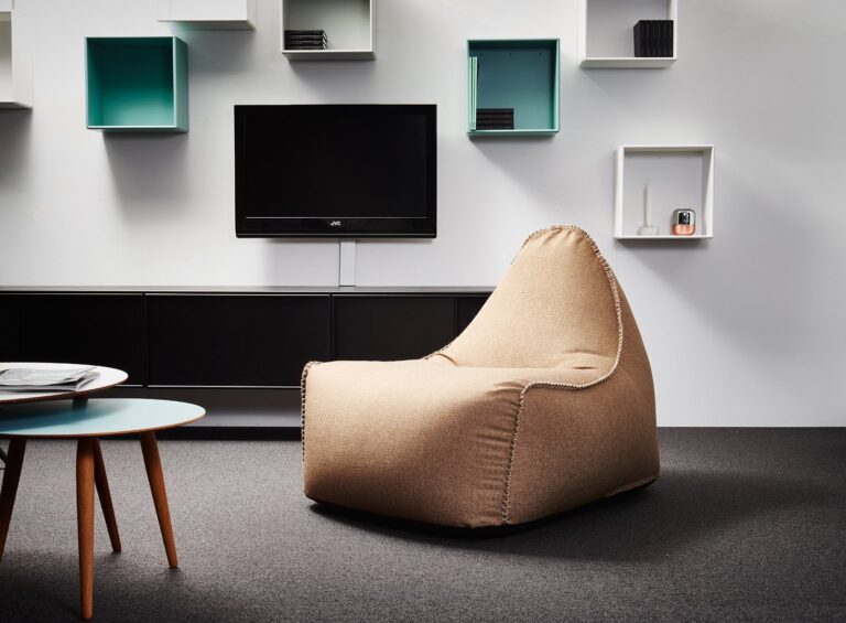 A beige bean bag chair sits in a modern living room with a wall-mounted telly, floating shelves, and a round coffee table.