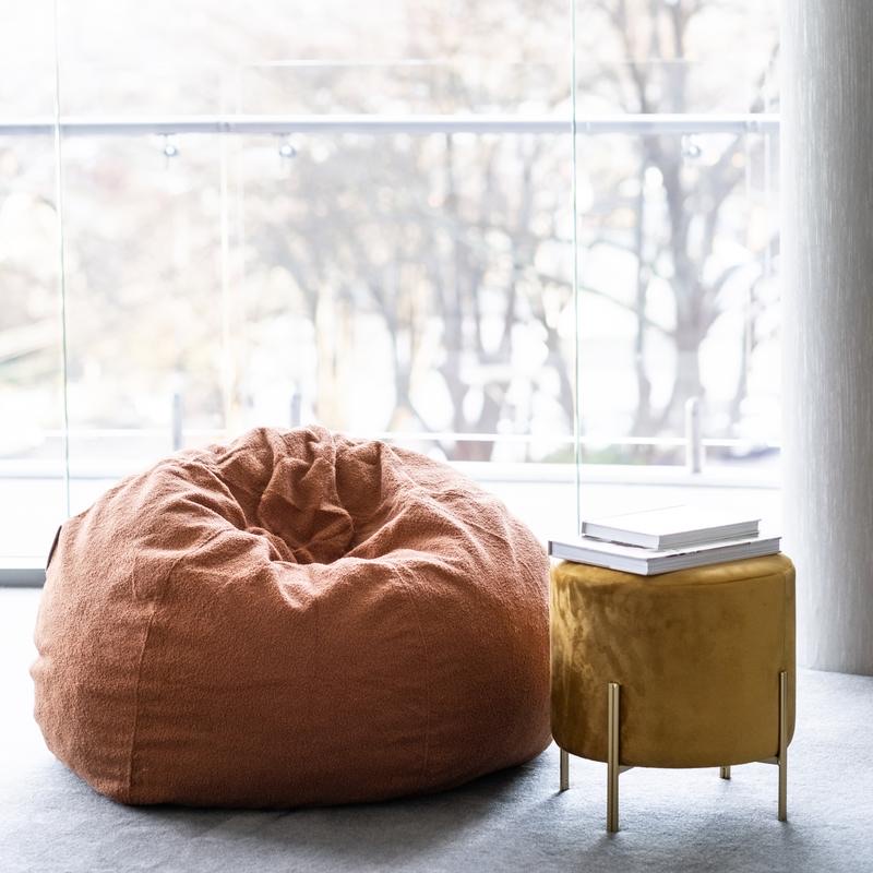 A brown beanbag sits next to a small yellow pouffe with books on top, in front of large windows overlooking trees.