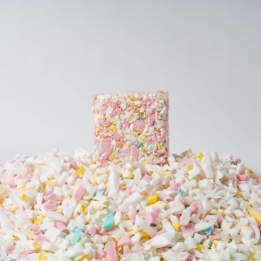 A rectangular pastel-coloured cereal treat stands upright among scattered pieces of marshmallow and cereal against a plain white background.