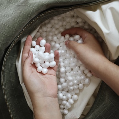 Two hands hold and sift through small white plastic pellets inside an open fabric container, possibly inspecting stuffing material.