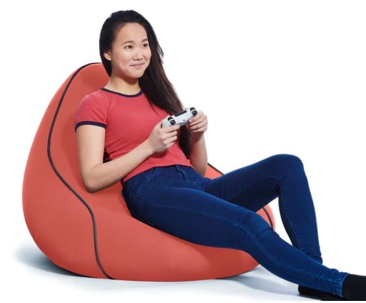 A person in a red shirt and blue jeans sits on an orange bean bag, holding a video game controller and looking ahead.