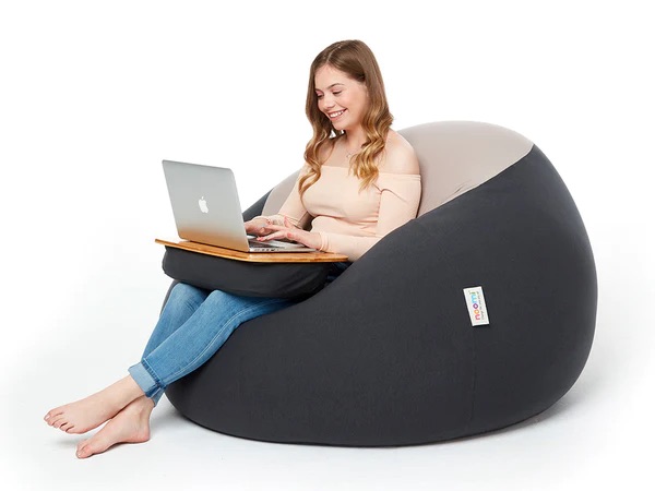 A person sits on a large black bean bag, using a laptop on a wooden lap desk, against a plain white background.