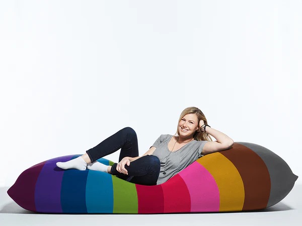 A woman reclines on a large, striped, bean-shaped cushion with colours including purple, blue, green, yellow, orange, and brown.