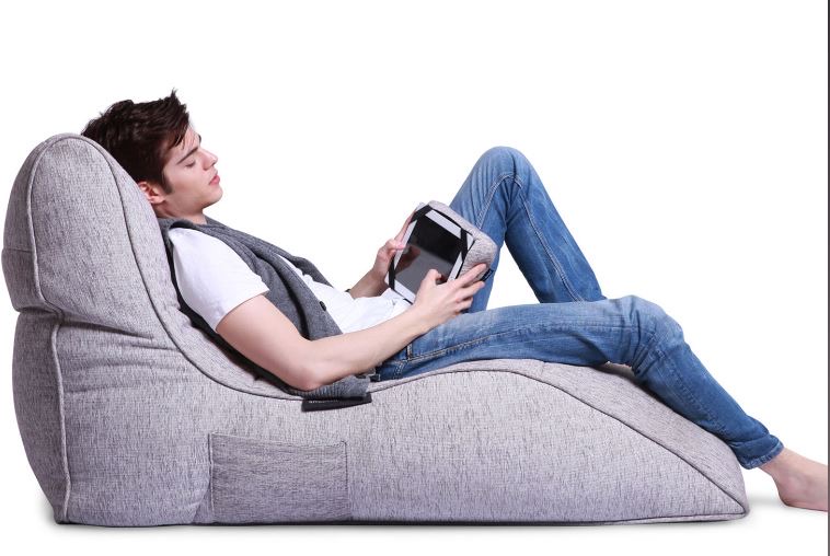A person is reclining on a grey bean bag, holding a tablet, wearing blue jeans and a white T-shirt against a plain white background.