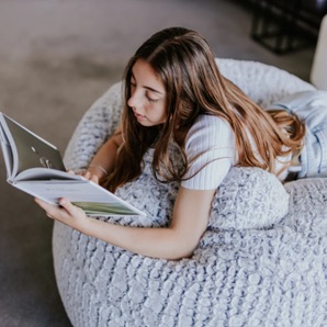 A young person with long hair is lying on a textured bean bag, reading an open book in a bright indoor setting.