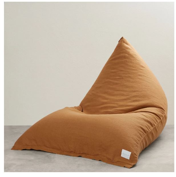 A large, triangular, brown bean bag sits on a grey floor against a plain off-white wall.