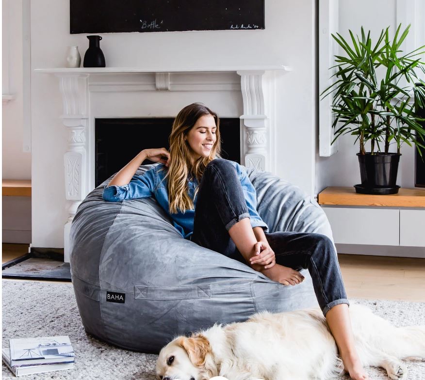 Woman sitting on a grey bean bag chair in a lounge, smiling, with a dog lying on the rug nearby.