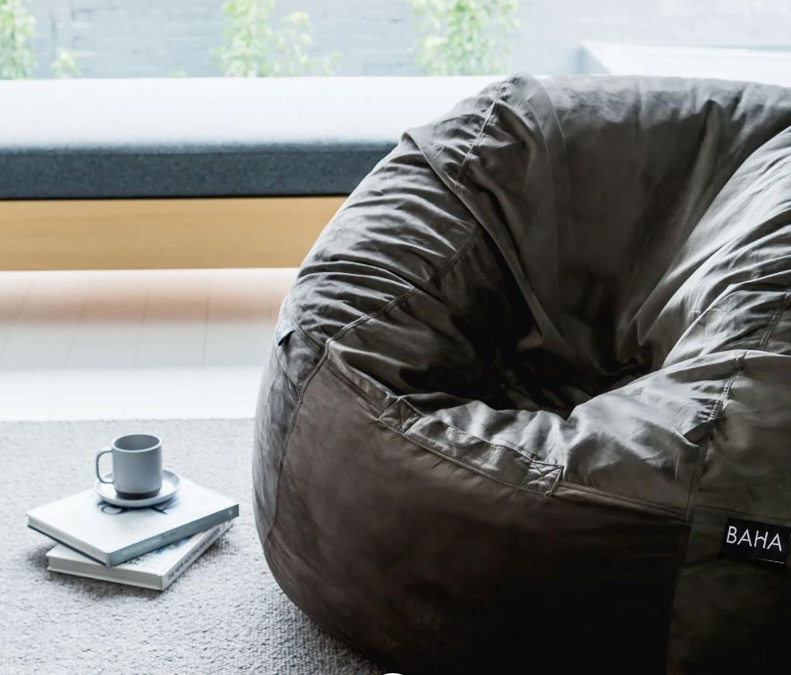 A large dark bean bag sits on a carpeted floor next to a stack of books and a cup and saucer, with a bench and window in the background.