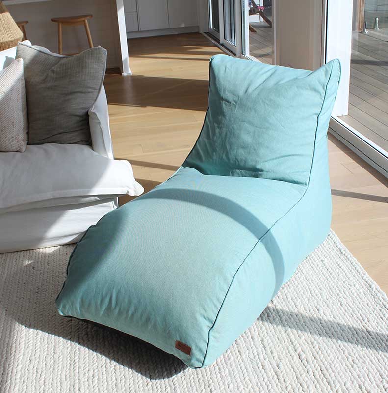 A light blue bean bag lounger sits on a textured white rug in a sunlit lounge near a white sofa and glass doors.