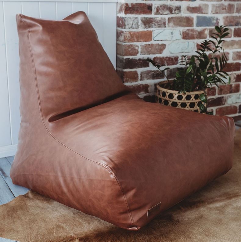 A brown leather beanbag chair sits on a brown rug near a brick wall and a potted plant.