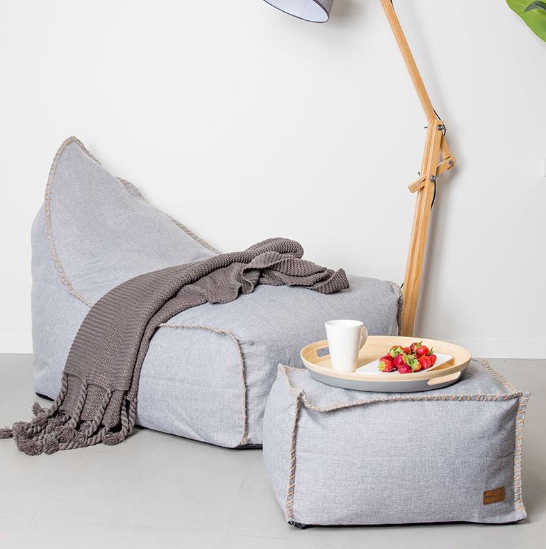 Grey bean bag chair with a grey knit throw, wooden floor lamp, and tray with a mug and strawberries on a matching ottoman, set against a white wall and light floor.