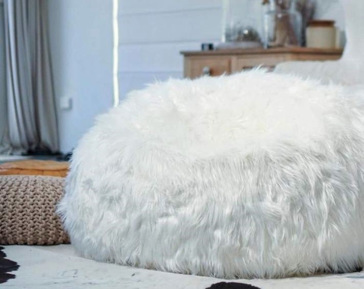A large, round, white faux fur bean bag sits on a bed in a cosy, softly lit room with neutral decor.