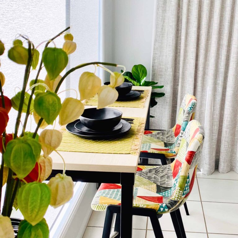A dining table with two colourful chairs, black crockery, a yellow placemat, and green leafy plants near a window with light curtains.
