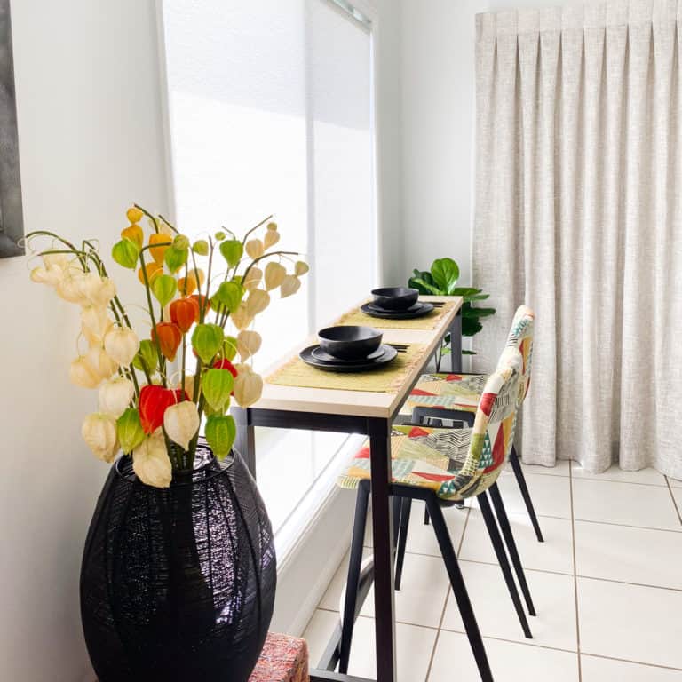 A small dining table set for two with black crockery, two colourful chairs, a tall vase with orange and white flowers, and a plant in the corner by a window with white curtains.
