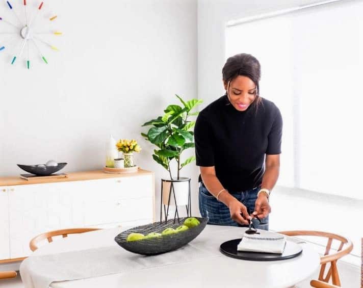 Woman standing at a dining table, stacking items on a tray in a bright, modern room with a plant, wall clock, and fruit bowl visible.