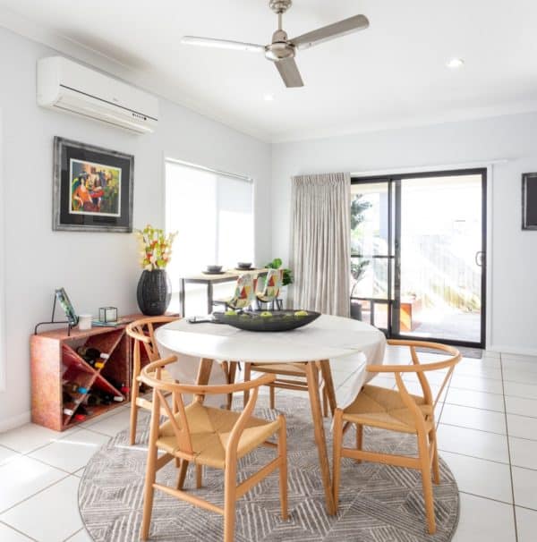 Bright dining area with a round table, four wooden chairs, wall art, wine rack, and sliding glass door leading outside. Ceiling fan and air conditioner are visible.