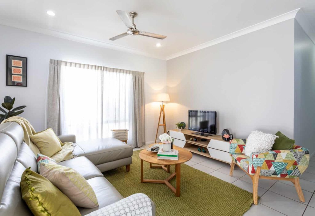 Modern living room with a grey corner sofa, patterned armchair, round wooden coffee table, TV unit, standard lamp, and green rug on tiled floor. Light enters through sheer curtains.